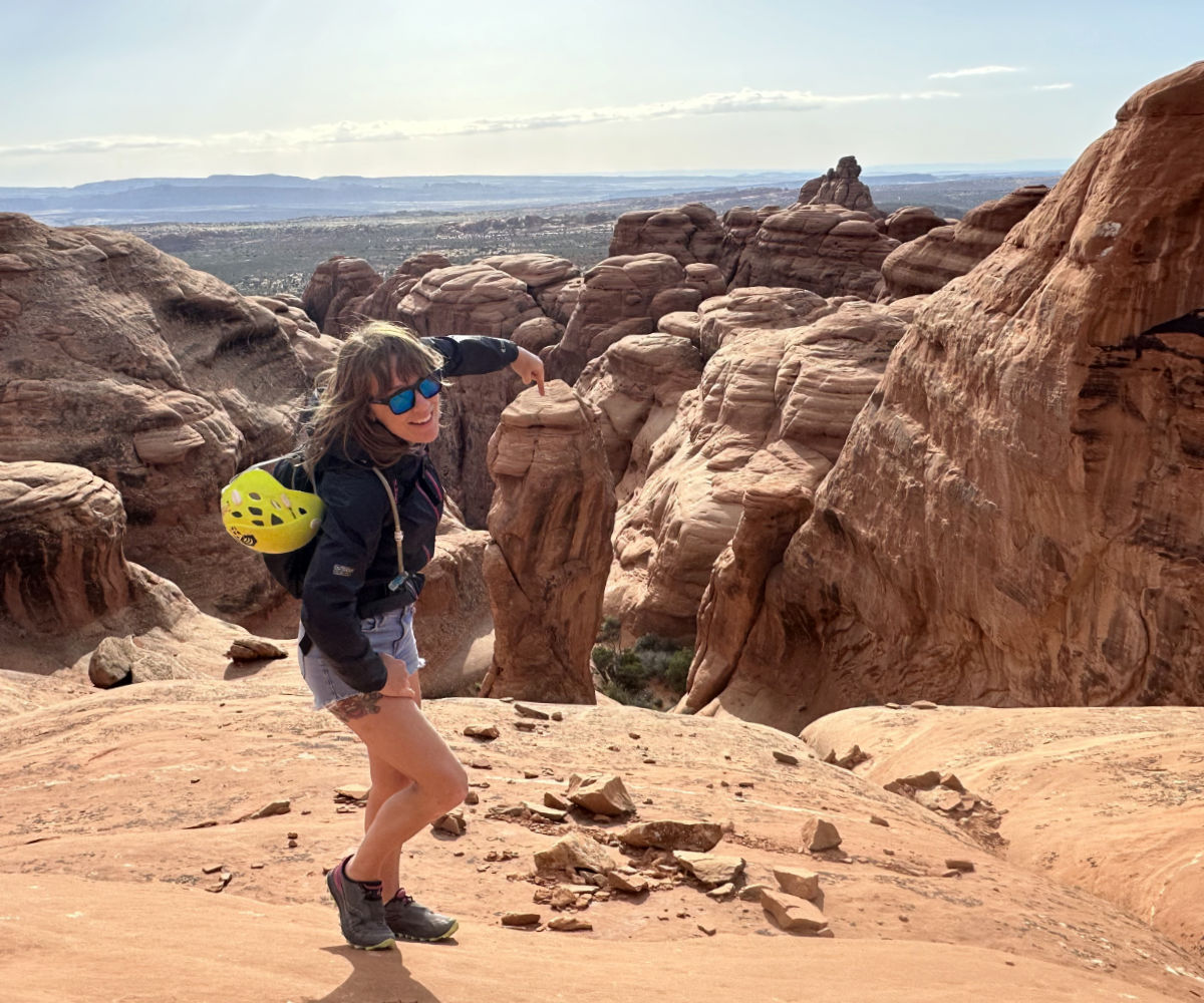 woman wearing jean shorts and windbreaker is pointing to sandstone tower at Arches National Park. Helmet hung on her backpack. Wispy clouds in the background